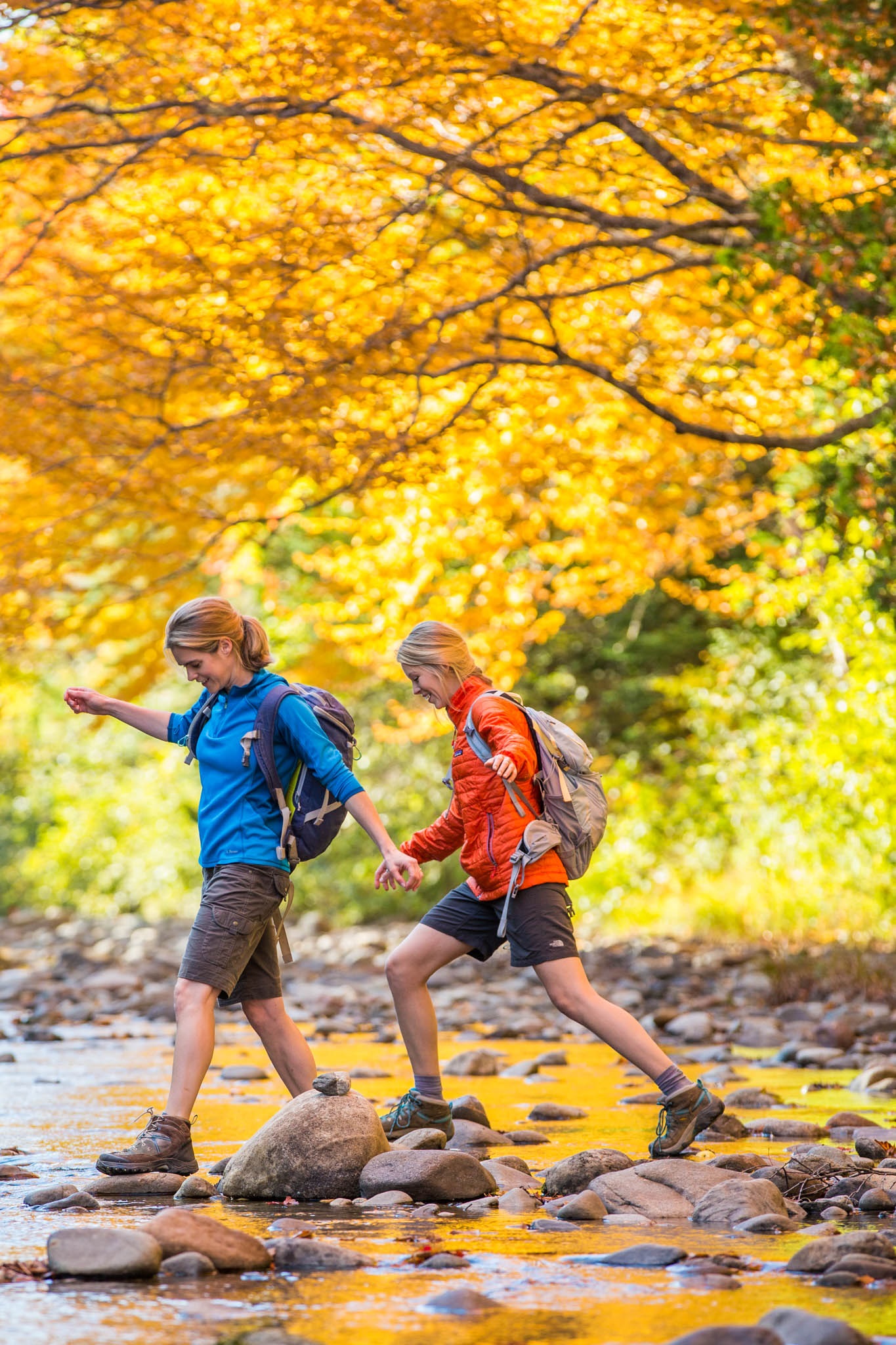 Two ladies hiking over a shallow stream in autumn.
